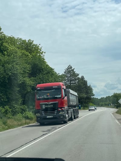 A photograph taken on a roadside in Tsar Kaloyan, Bulgaria, shows a red MAN truck driving on a gray asphalt road, with lush green trees lining the road's edge on both sides. A white vehicle is visible further down the road. The sky overhead is cloudy. The truck's license plate is visible as P 6276 MB.