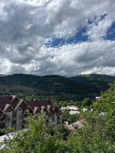 The image captures a panoramic view of the city of Dilijan, Armenia, under a dramatic sky filled with a mix of dark and light clouds. In the foreground, a large, multi-story building with a distinctive red, triangular roof design dominates the lower left of the frame. Several balconies with railings are visible along its facade. To the right and in front of this building, lush green trees with abundant foliage partially obscure the view of other buildings and houses.

Behind the foreground structures, the landscape transitions into rolling hills covered in dense green forests. The trees vary in shade, indicating different types of vegetation. In the distance, larger mountains rise, their peaks softened by the atmospheric haze and the cloudy sky. The sky itself features patches of bright blue interspersed with thick, grey clouds, suggesting a dynamic weather system. A distant transmission tower can be seen on one of the hills. The overall impression is one of a town nestled within a verdant, mountainous region.