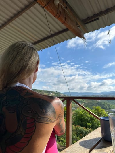 A person with blonde hair and a large tattoo of a snake on their back is seen from behind, looking out at a lush green landscape with rolling hills and scattered buildings under a bright blue sky with white clouds. They are sitting at a wooden table on a covered porch, with a blue mug and a clear glass in front of them. The porch has a wooden railing. The scene is set outdoors, likely in a tropical or subtropical environment, with the architecture suggesting a rustic setting, possibly in or near Thôn 3, Vietnam. The person appears relaxed, enjoying the view.