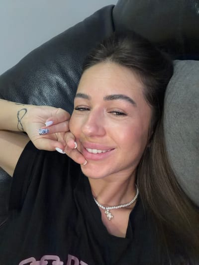 A close-up, eye-level, selfie-style shot captures a woman lying down, resting her head on a dark leather surface and a grey cushion. She is looking towards the camera with a slight smile, her right hand touching her cheek. Her left arm is visible, featuring an infinity tattoo on the wrist and manicured fingernails with a white and sparkly design. She is wearing a black t-shirt and a pearl necklace with a diamond-encrusted teddy bear pendant. The background is indistinct, suggesting an indoor setting. The image does not contain any information about the city of Novi Iskar, Bulgaria.