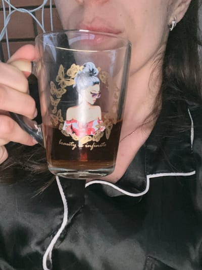 A close-up shot captures a person holding a transparent glass mug filled with a dark beverage. The mug features a graphic illustration of a woman with a bun hairstyle, wearing sunglasses and a pink and white top, surrounded by golden butterflies. Below the illustration, the words "beauty is infinite" are inscribed in gold. The person is wearing a black satin pajama top with white piping. The background appears to be an outdoor setting with a blurred brick wall and a light blue wire fence. The image is well-lit, with the mug being the central focus. The person's hand is visible, holding the mug's handle, and part of their face, including their lips and nose, is in view. A silver earring is visible on the person's right ear. The overall scene suggests a relaxed moment, possibly in the morning or evening. The image does not provide any specific details or indicators that would allow for identification of a location in Kaspiysk, Russia.