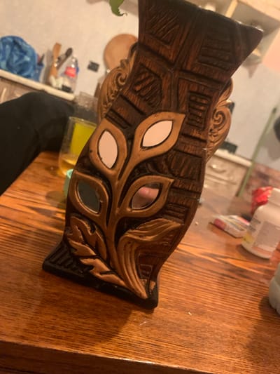 A close-up, medium-shot photograph captures a decorative wooden vase with carved leaf and geometric patterns, placed on a wooden table. The vase is a deep brown with lighter brown carved accents, featuring reflective white and dark mirrored inserts within the leaf designs. In the blurred background, a yellow liquid in a glass and a black fabric object are visible. To the right, a white plastic bottle and a box of medication are also out of focus. The scene is indoors, suggesting a home environment. The photograph does not contain any elements that would allow for the determination of its location in Krushev Dol, Bulgaria.