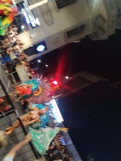 A tilted view captures a vibrant street parade at night. Dancers in ornate, colorful costumes with feathers and sequins are the focal point. Crowds line the street, with one person's hand partially visible in the foreground. Above, blurred signs of a "Coffee Lounge" and a Starbucks logo can be seen. Red lights from traffic or building illumination pierce the darkness above the parade.
