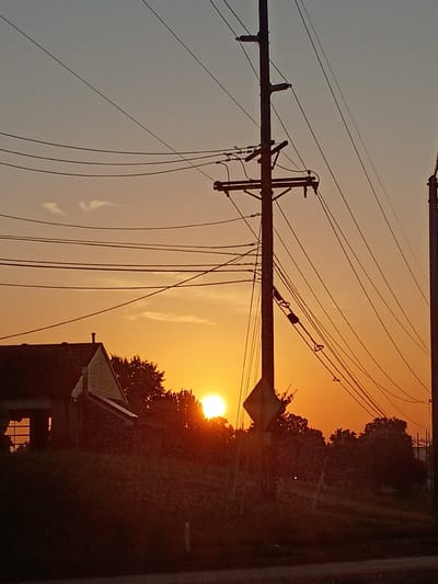 The image captures a sunset in a rural or suburban setting. The sun is low on the horizon, casting a warm, orange glow across the sky and silhouetting trees and structures. Power lines crisscross the frame, adding a sense of urban infrastructure to the natural beauty of the sunset.
The main subjects include utility poles with numerous wires, a portion of a building with a peaked roof, and the natural elements of trees and the sky. The sun itself is a prominent subject, with its bright orb partially obscured by foliage. The scene is dominated by the warm hues of the sunset, ranging from pale yellow to deep orange and red.
The activity taking place is passive; it's a moment of natural beauty as the day ends. The time of day is clearly sunset, indicated by the low angle of the sun and the dramatic lighting. The weather appears to be clear, allowing for an unobstructed view of the sunset, though some wispy clouds are visible in the sky.
Notable details include the intricate network of power lines and the silhouetted structure that appears to be a house or a small building, with a window visible. The overall emotion evoked is one of tranquility and the natural beauty of a day's end. There is no visible text in the image. The context provided indicates the location as Frankfort, United States of America.