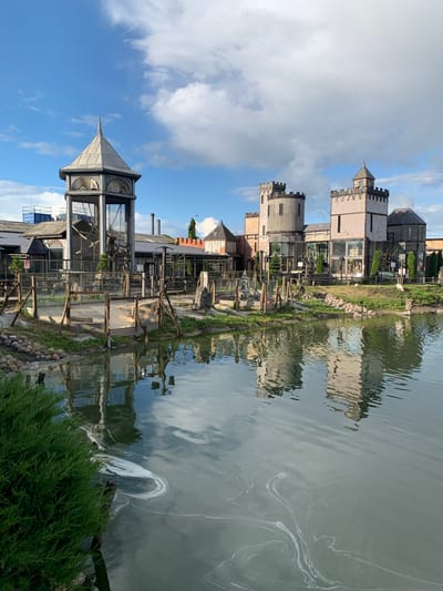The image shows a daylight, exterior view of an amusement park or attraction featuring a castle-like structure and a water feature. The main structure is a series of interconnected buildings with towers and crenellations, resembling a medieval fortress. One prominent tower has a pointed, conical roof. The buildings appear to be constructed with a mix of materials, including stone, brick, and glass. In front of the castle structure is a body of water, possibly a pond or small lake, with reflections of the buildings visible on its surface. There are also some wooden fences and enclosures in the foreground, suggesting animal exhibits or play areas. Greenery, including trees and bushes, is present around the water and the structures. The sky is blue with scattered white clouds. The overall scene suggests a recreational or entertainment venue designed with a historical or fantasy theme. The provided location "Малахавецкі сельскі Савет, Belarus" indicates this is a rural area in Belarus.