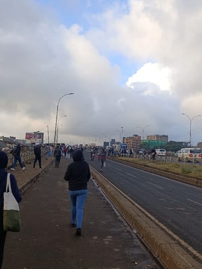 A street scene unfolds beneath a cloudy sky, with a glimpse of blue peeking through. Pedestrians stroll along a wide walkway, while vehicles traverse the adjacent road. Streetlights line the route, and buildings are visible in the distance. The overall atmosphere suggests an urban environment on a somewhat overcast day.