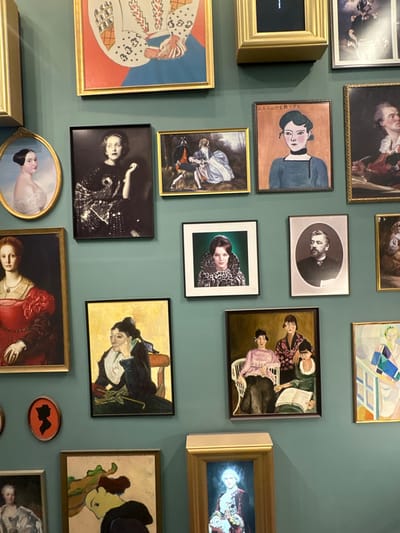The image displays a gallery-style arrangement of multiple framed artworks on a dark teal or green-blue wall, likely located in Levallois-Perret, France. The collection includes various styles and periods, predominantly portraits.
Notable artworks include:
* An oval-framed classical portrait of a fair-skinned woman with dark hair, shoulders exposed.
* A large rectangular gold-framed portrait of a woman in a red dress, in a classical style.
* A black-framed black and white photograph of a woman in a sparkling, patterned garment, posing with one hand near her face.
* A gold-framed painting depicting two figures in historical attire within a landscape, one seated, one kneeling.
* A black-framed portrait of a woman with a dark bob haircut and a choker, labeled "MARGUERITE," against an orange background.
* A square black-framed color portrait of a woman with dark hair wearing a leopard print collar.
* A silver-framed black and white portrait photograph of a bearded man in formal attire.
* A black-framed painting of a seated woman, head resting on her hand, in a dark dress.
* A black-framed painting depicting three women, two seated on a white wicker chair, one reading a book, another standing behind them.
* A round, orange and black silhouette profile of a woman.
Various other framed pieces, including abstract, modern, and classical portraits, are partially visible, creating a dense display. The frames vary in material and style, including gold, black, and silver tones. No people are present within the observable scene, only depicted in the artworks.