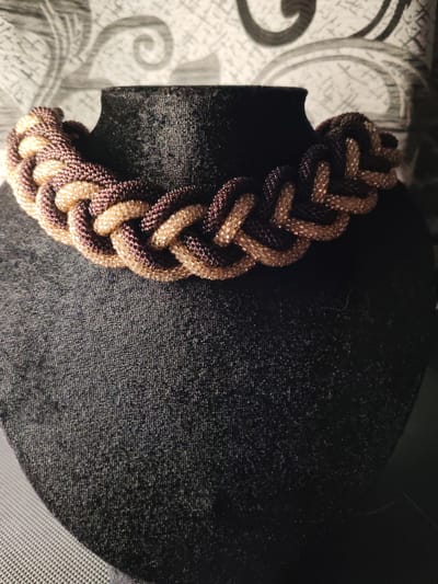 A braided necklace made of small beads in shades of brown and tan is displayed on a black velvet bust. The necklace is intricately woven, forming a rope-like design. The bust is positioned in front of a textured, grey and white patterned wallpaper. The background is dimly lit, with some areas in shadow. The lower part of the image shows a dark, textured surface, possibly the base of the display. The image was taken indoors. There is no indication of the location being in Сокол, Russia.