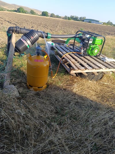 In Kurtovo Konare, Bulgaria, an outdoor scene depicts agricultural equipment set up in a dry, grassy field. A yellow propane tank stands upright on the ground, connected by an orange hose to a green and black engine-powered water pump. The pump is positioned on a wooden pallet, which rests on a black tarp or container. A black filter is attached to a black pipe, supported by a wooden stake. A green hose extends from the pump, presumably for water distribution. In the background, a tilled field stretches towards rolling hills dotted with trees and buildings, under a clear blue sky. The lighting suggests it is daytime.