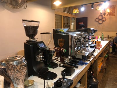 The image shows the interior of a coffee shop, likely in Tambonbaroh, Indonesia. The scene is dominated by professional coffee-making equipment. On the left, a large black coffee grinder with a clear hopper full of coffee beans is prominent. To its right, a sleek, chrome espresso machine with multiple levers and gauges sits on a wooden counter. Various coffee-making tools, such as portafilters and a tamper, are neatly arranged on the counter. Bags of coffee beans labeled "Pusrain Coffee" are visible in the foreground, suggesting a focus on freshly roasted coffee.
In the background, a person's head is visible through a doorway, possibly a staff member. The setting appears to be a brightly lit indoor space, with warm lighting from overhead lamps and a decorative chandelier. The walls are a pale color, and there are wooden shelves built into the counter, displaying some beverage cartons. The overall atmosphere suggests a functional and well-equipped coffee preparation area, possibly during operational hours. There is no indication of the time of day or weather conditions outside. The visible text includes "Pusrain Coffee" on the coffee bags, and some smaller text on a beverage carton that is not fully legible. The scene conveys a sense of professionalism and dedication to coffee craft.