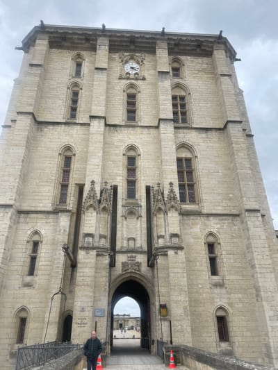 The image displays the imposing Donjon (Keep) of the Château de Vincennes, located in Vincennes, France. The central subject is a tall, light-colored stone tower, exhibiting medieval fortress architecture with Gothic influences. Its facade features numerous narrow, arched windows with dark panes. A prominent clock face, showing approximately 10:45, is centered on an upper section of the tower. Below, a large arched gateway serves as the main entrance, leading into a shaded passage and an exterior courtyard where distant figures are visible. Ornate stone carvings are positioned above the archway. Flanking the entrance are deep vertical recesses, suggesting former drawbridge channels, with heavy chains visible descending on the left side.
In the foreground, a stone bridge with dark metal railings extends towards the tower's entrance. On the bridge's left side, near the railing, a sign reads "VISITE CHATEAU." One adult male, wearing a dark jacket, stands on the bridge structure to the left of the entrance, facing slightly towards the archway. Two orange traffic cones are present on the bridge, one on each side of the central path. The sky is overcast and grey, providing soft, diffused lighting to the scene.