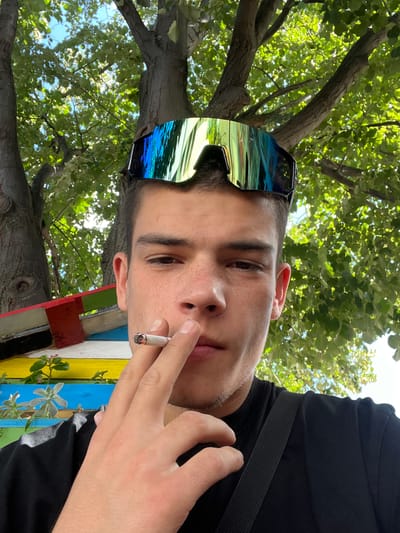 In a medium shot, a young adult male is captured from the chest up, outdoors. He is wearing a black t-shirt and large, reflective goggles pushed up onto his forehead. His right hand is raised to his mouth, holding a lit cigarette between his index and middle fingers. A portion of a colorful, multi-colored structure is visible behind him, with red, yellow, blue, and green sections. The background is dominated by the lush green foliage of trees and a bright blue sky. The image appears to be a selfie, taken in natural daylight. The specific location of Sopot, Bulgaria is not discernible from the visual elements of the photograph.