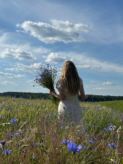 A woman with long, blonde hair, viewed from behind, stands in a field of green wheat and blue wildflowers under a blue sky with white clouds. She wears a white, short-sleeved dress with a subtle pattern and holds a large bouquet of blue wildflowers. The field extends to a tree line in the background. The scene is located in the territory of Parytski Sel'ski Sovet, Belarus.
