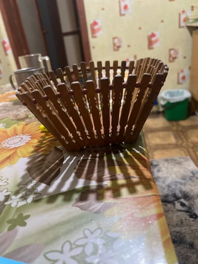 An indoor scene shows a wooden, expandable basket on a table covered with a floral-patterned tablecloth. The basket is made of vertical wooden slats connected by small red beads. A shadow from the basket is cast on the tablecloth. In the background, a metal mug sits on the table. The wall behind the table has a yellow wallpaper with a repeating pattern of framed images. To the right, a green trash can with a white liner is visible. The flooring appears to be tiled. The image is taken in Luhansk, Ukraine.