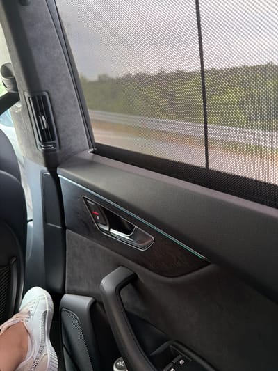 The image depicts the interior of a car, viewed from the perspective of a passenger. The scene focuses on the rear door panel, which is finished in dark leather and features a wood-grain accent panel. A chrome door handle with a red lock indicator is visible, along with a thin LED light strip highlighting the trim. The window is partially covered by a mesh sunshade, through which a blurred landscape of trees and a road can be seen. In the foreground, a person's foot wearing a white sneaker is resting on the car floor. The car's interior includes air vents and a speaker grille. The overall impression is one of a typical car interior, likely on a journey. The location is specified as Balgarchevo, Bulgaria, suggesting the image was taken there.