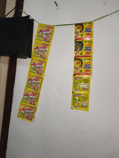In the city of Barengkrajan, Indonesia, a simple wall serves as a makeshift display for two vertical strings of seasoning packets. On the left, a string of "Masako" brand seasoning packets, specifically the "Daging Sapi" (beef) flavor, hangs in a descending line. The packets are predominantly yellow with red and green accents, featuring images of beef broth and ingredients. On the right, another string holds "Magic Lezat" seasoning packets, also predominantly yellow with red and white lettering, displaying images of cooked dishes. These packets appear to be promotions, indicated by text like "BELI 10 GRATIS 1" (Buy 10 Get 1 Free). Above and to the left of the seasoning packets, a black bag with a textured surface is suspended by purple and green cords, adding a touch of domesticity to the scene. The wall itself is white and textured, with a few minor scuffs and smudges, suggesting a well-used living space. The lighting is dim, casting subtle shadows and highlighting the texture of the wall. A single screw is visible on the wall to the upper right, possibly a remnant of a previous hanging or decoration.
