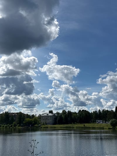 The image depicts a partially overcast sky over a tranquil body of water. The sky is a vibrant blue with scattered cumulus clouds, some of which are dark and imposing, while others are bright and fluffy, catching the sunlight. The clouds are varied in shape and size, creating a dynamic and somewhat dramatic sky.

On the right side of the image, the water stretches out, appearing still with subtle ripples reflecting the sky and the trees along the bank. The reflections are slightly distorted by the movement of the water.

Along the bank of the water, a dense line of trees forms a verdant border. Behind the trees, several buildings are visible. One building in the distance appears to be a multi-story structure with a light-colored facade and visible windows. Closer to the foreground, another building with a darker roof is partially obscured by trees. The presence of these buildings suggests an inhabited area, possibly a town or a residential complex situated near the water.

The overall scene evokes a sense of natural beauty combined with human presence. The lighting suggests it is daytime, likely late morning or early afternoon, given the brightness of the sky and the illumination of the clouds. The weather appears to be a mix of sun and clouds, creating a visually interesting contrast. The reflection of the sky and greenery on the water adds depth and serenity to the image. There is no visible text in the image.