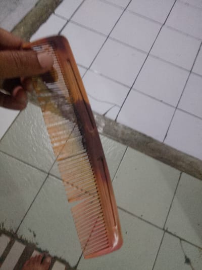 A person's hand is holding a comb with brown and orange tones. The comb has fine teeth and a slightly curved handle. The background appears to be a tiled floor with a grid pattern. The tiles are light-colored, possibly white or off-white, and are separated by darker grout lines. In the upper portion of the image, there are also some darker, irregular tiles or stones. A person's bare foot is visible in the bottom left corner of the image, with toes and a heel showing. The scene is likely indoors, given the tiled floor, and the lighting suggests it could be daytime. There is no visible text in the image. The location context is Cisarua, Indonesia, which suggests a tropical or subtropical climate, though this cannot be definitively determined from the image alone. The image is somewhat grainy and out of focus, particularly in the background.