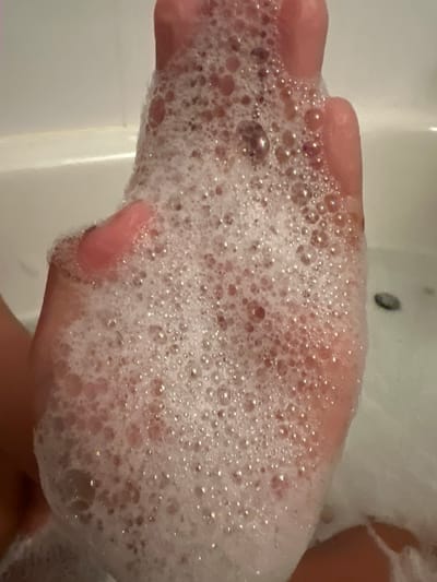 A close-up, overhead shot shows a hand submerged in bath bubbles. The hand is pinkish and appears to be either a child's or a woman's. The bubbles are white and foamy, covering most of the hand and extending out of frame. Some of the bubbles have a shimmering quality, suggesting light reflecting off them. In the background, a portion of a white bathtub is visible. The scene is likely set in a residential bathroom. There is no identifiable location in Surrey, Canada.