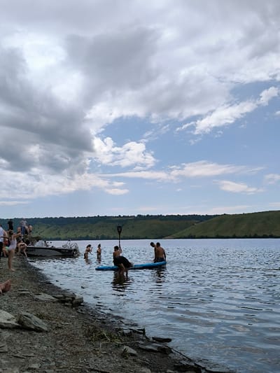 This image captures a daytime scene on the Dniester River near Horaivka, Ukraine. The sky is partially overcast, with dense grey clouds dominating the left and upper-center, transitioning to a clear blue sky with scattered white clouds on the right. The foreground consists of a rocky and gravelly shoreline that gradually slopes into the water.
Approximately 15-20 individuals are present in the scene. On the left side of the shoreline, a group of people are gathered, some standing and others partially obscured or seated on the rocks. A grey and black motorboat, bearing the visible registration "UMS-470", is partially beached on the left bank, adjacent to this group.
In the mid-ground water, at least seven individuals are visible. Centrally, two shirtless males are standing waist-deep next to a blue stand-up paddleboard (SUP); one of them holds a black paddle, while another individual is partially submerged and interacting with the SUP. To their left, a female in a dark two-piece swimsuit and another male are standing in the water. Other individuals are further back in the water, less distinctly visible. The water itself is calm with minor ripples.
The background is dominated by a continuous stretch of green, undulating hills covered in vegetation, extending across the horizon and bordering the wide river.