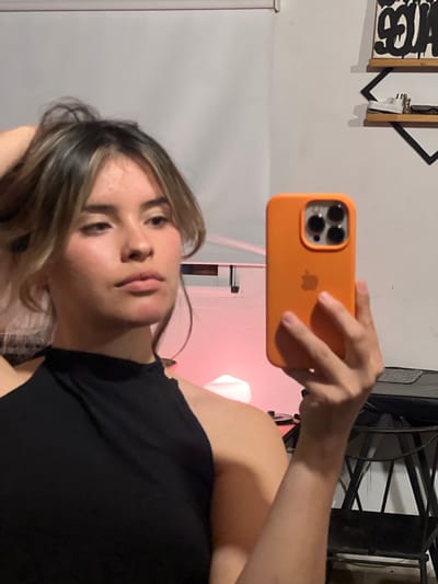 A person is taking a selfie with an orange-cased iPhone, indoors. The background includes a white wall with decorative elements, a lamp emitting a pink hue, and a black metal structure. The person is wearing a black top. The location is Sumalao, Argentina.