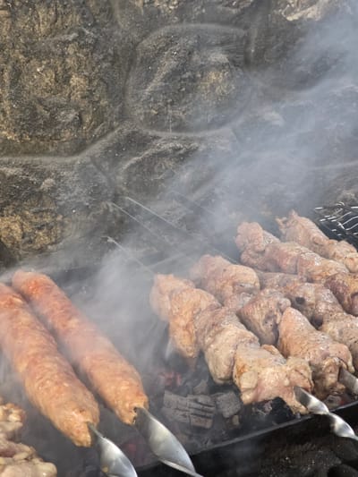 This media file shows a close-up of skewers of meat being cooked over hot coals, with smoke rising. The scene appears to be outdoors, with a rough, textured surface to the left of the food, possibly a stone wall. The lighting suggests it is daytime.
The primary subjects are skewers of meat. Some skewers hold chunks of meat, likely pork or beef, which are seasoned and appear to be in the process of grilling. Other skewers hold elongated, sausage-like meat, which are also cooking. The meat is a light brown color, indicating it is cooked or cooking. There are visible signs of charring on some pieces. The cooking apparatus includes metal skewers and what appears to be a metal grill grate. Hot coals are visible beneath the meat, providing the heat source, and smoke is billowing around the food, creating a hazy effect.
The activity is clearly the cooking of meat on skewers, a common method for preparing kebabs or shashlik. The event is likely a barbecue or outdoor meal preparation.
Notable details include the texture of the stone-like surface to the left, which is rough and uneven, with hints of tan and dark grey. The smoke is thick and obscures some of the details of the grilling area, adding to the atmosphere of cooking. The metallic sheen of the skewers is also noticeable. No people are visible in the media file. There is no visible text. The location context provided is Pidhorodne, Ukraine, suggesting a rural or suburban setting where outdoor cooking might be common.