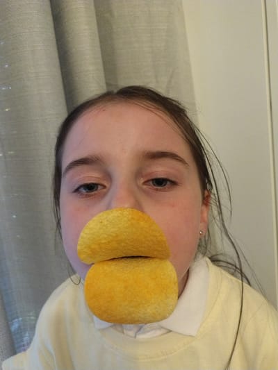 A young person's face is partially obscured by two potato chips placed over their mouth, resembling a beak. The person has fair skin, blue eyes, and dark brown hair that is pulled back. They are wearing a light yellow sweater over a white collared shirt. The background consists of a light-colored curtain. The scene is likely indoors, possibly in a residential setting. There is no identifiable information within the image to suggest a specific location in Crauthem, Luxembourg.