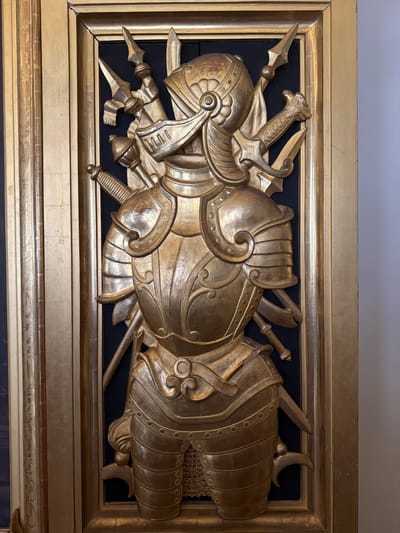 This image presents a vertically oriented, gilded three-dimensional relief depicting a partial suit of armor and weaponry, an object located in Città del Vaticano, Vatican City. The central subject is a highly detailed, golden-finished replica of a knight's cuirass and helmet, crafted with visible segmented plates, decorative embossing, and rivet details. A segment of chainmail is discernible beneath the lower portion of the armor. Behind the central armor piece, an array of stylized golden weapons is arranged, including spearheads, a mace-like weapon, and several swords, one featuring an ornate hilt shaped like a beast's head. The entire golden relief is set against a dark, matte black background, providing strong visual contrast. This panel is encased within a robust gilded wooden frame; the inner frame is simpler, while the broader outer frame on the left exhibits intricate carved scrollwork and vertical fluting. To the immediate right of the frame, a plain, light-colored wall is visible, suggesting an interior setting. On the far left, additional gilded architectural elements or paneling are partially visible. The lighting illuminates the golden surfaces, emphasizing their metallic luster and three-dimensional contours.