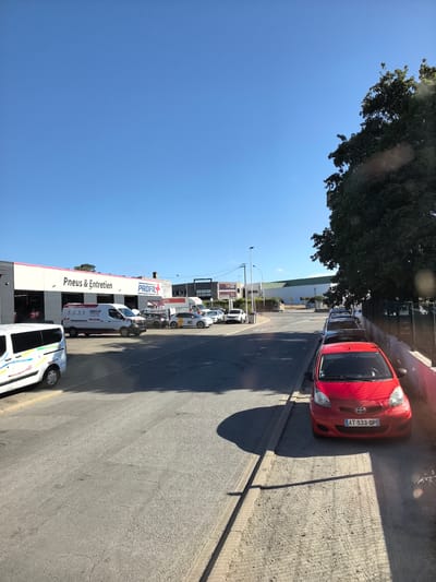 The image shows a commercial area in Saint-Herblain, France, likely a business park or industrial zone. In the foreground, a red Toyota Aygo with the license plate AT-533-QP is parked on a dirt patch next to a paved road. The car is facing forward. To the left, a white van with colorful decals is partially visible. Behind the van, a building with a pink and white facade is labeled "Pneus & Entretien" and "PROFIL". Several cars are parked in front of this building. Further down the street, more buildings and vehicles are present, including a white truck. A large, leafy tree is on the right side of the image, casting a shadow on the ground. The sky is clear blue.