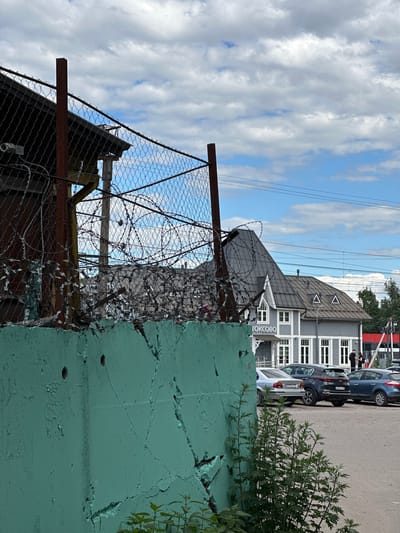 The image shows a view of the Toksovo train station in Russia. In the foreground, a weathered, light green concrete wall runs across the bottom of the frame, with some green plants growing near its base. A tall metal fence with barbed wire runs horizontally above the wall. Behind the fence, a two-story, gray wooden building with white trim, identified by a sign reading "ТОКСОВО" (Toksovo), stands as the train station. The building has a steep, dark gray roof with dormer windows. A parking lot with several cars is visible in front of the station. The sky is partly cloudy with blue patches. A few people are standing near the entrance of the train station.