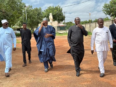 Six men are walking on a reddish-brown dirt path with gravel. The man on the far left is wearing a light blue traditional outfit and a white cap. Next to him is a young man in a black traditional outfit. The third man from the left is wearing a dark blue traditional outfit and a patterned cap, and he is holding a phone to his ear. The fourth man from the left is wearing a black traditional outfit and glasses, and he is touching his collar. The fifth man from the left is wearing a white traditional outfit. The sixth man from the left is wearing a dark suit and a red and white patterned shirt. Trees and utility poles are visible in the background, and the sky is cloudy. The location is identified as Kabala, Nigeria.