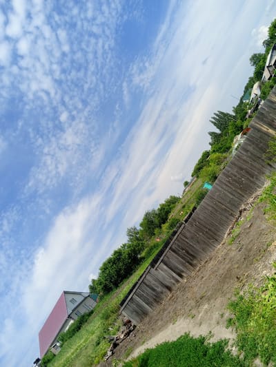 This is a tilted, outdoor, eye-level, full shot of a rural landscape in Pavlovsk, Russia. The sky is a bright blue with scattered white, wispy clouds. In the foreground, a weathered wooden fence runs diagonally across the frame. Beyond the fence, a grassy slope is covered in lush green vegetation. A white house with a red-tiled roof is visible on the left side of the frame, nestled among the greenery. Further in the distance, more houses and trees can be seen, suggesting a residential area. The overall scene evokes a peaceful, summer day. The lighting suggests it is daytime, likely late morning or early afternoon, with the sun casting soft shadows. There are no people visible in the image, and no text is present.