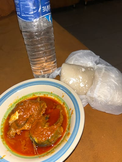 This image depicts a meal served on a brown surface, presumably a table, in an indoor setting. The scene is stated to be located in Egbe, Nigeria. The primary focus is a white bowl with blue and green striped rims containing a reddish-orange stew with two pieces of fish, likely catfish, visible within the sauce. Adjacent to the bowl are two portions of a light-colored, solid food item, consistent with a 'swallow,' individually wrapped in clear plastic bags. A transparent plastic bottle of "DAVIE" brand drinking water, partially consumed, stands upright behind the food items. No people or interactions are visible.