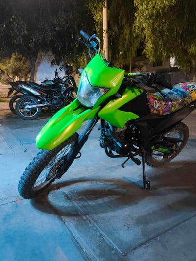 The image shows two motorcycles parked on a paved surface, likely a sidewalk, in an urban environment. The foreground motorcycle is a bright green dual-sport or off-road type. It has a distinctive custom seat cover with cartoonish graffiti-style graphics. The motorcycle is resting on its kickstand. Behind it, there is another motorcycle, dark in color, with red accents, parked parallel to the green one. The background includes trees and buildings, suggesting a city street at night. The image was taken in Trujillo, Peru.