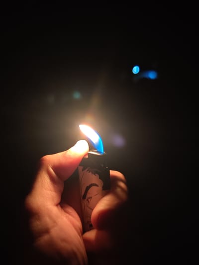 A human hand, appearing adult, holds a black plastic lighter that emits a vibrant blue flame with an orange base. The flame intensely illuminates the skin of the hand, casting a warm glow. The side of the lighter features a black and white stylized illustration of a male figure's profile, characterized by a distinct dark eyebrow and part of an eye. The background is predominantly dark, suggesting an unlit environment. In the upper right quadrant of the background, two out-of-focus blue light sources are visible, presenting as distinct bokeh effects. A fainter, diffused light source with an amber hue is present in the lower background. The image provides no visual information to confirm the location as Veliki Preslav, Bulgaria.