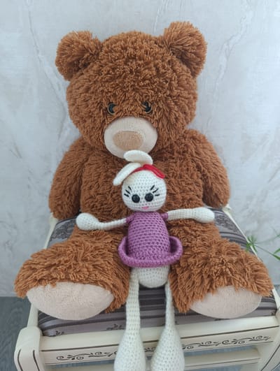 A plush brown teddy bear sits on a chair with a small, crocheted white rabbit in its lap. The teddy bear has a lighter brown muzzle and paws. The rabbit is wearing a purple dress and has a red bow on its head. It has black button eyes and a pink crocheted nose and mouth. The chair has a striped cushion and is made of white wood with decorative carvings. The background is a textured white wall. This scene is located in Pivdenne, Ukraine.