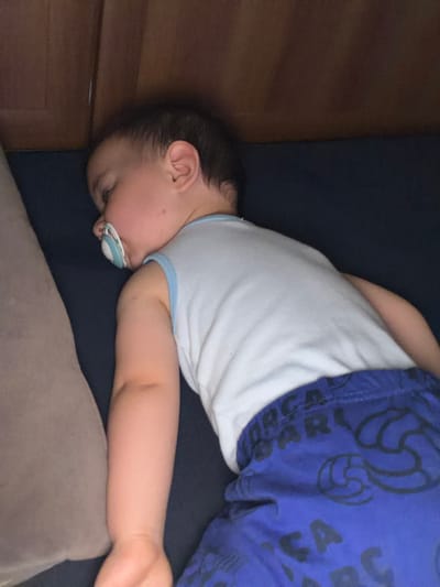 A baby is lying on their side, face down, with a pacifier in their mouth. The baby is wearing a white tank top with light blue trim and blue shorts with a pattern of black sports balls and letters. The baby's head is near a wooden corner, possibly of a crib or bed. The baby's arm is extended along a soft, light-brown pillow or cushion. The surface the baby is lying on appears to be dark blue. There is no contextual information in the image to suggest a location in Sevlievo, Bulgaria.