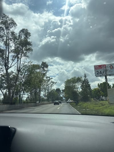 The image presents a forward-facing view from within a vehicle, likely a car, onto a multi-lane highway in Cholula, Mexico. The bottom quarter of the frame is occupied by a dark gray dashboard, with a dark, unidentified object partially visible on its left side. The paved road, marked with white lane lines, extends into the distance and carries moderate traffic, including several sedans and a white pickup truck with a ladder rack. Dense green vegetation, primarily tall, slender trees, borders both sides of the road. On the right, a concrete barrier separates the road from a grassy embankment and an area of disturbed earth. Above this, a large billboard stands prominently, displaying an advertisement for "TOYOTA" in red and white text, stating "EN TOYOTA SÍ" and "CONTAMOS CON ATENCIÓN 24/7" (We provide 24/7 service), accompanied by an image of a woman. The upper half of the image features a dynamic sky with large, dark grey cumulus clouds interspersed with patches of blue sky, through which distinct shafts of sunlight penetrate, particularly in the upper left quadrant. No human individuals are discernible in the actual scene, only the depicted person on the billboard.