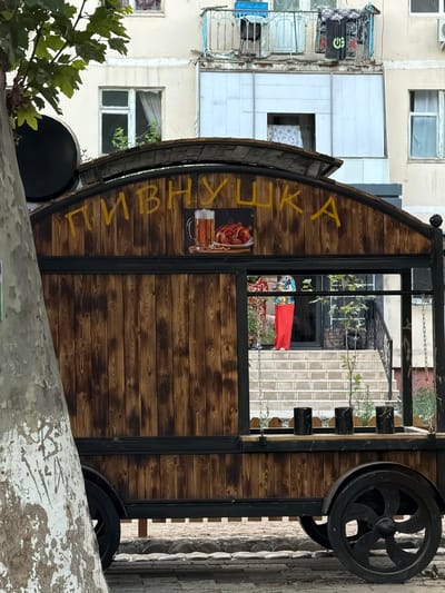 The image captures a detailed view of a rustic wooden kiosk, likely a food or drink stand, set against the backdrop of a building. The kiosk features a curved roof and is adorned with yellow Cyrillic lettering that appears to read "ПИВНУШКА," possibly indicating a beer-related establishment. A graphic of a beer mug and roasted chicken is prominently displayed on the front. The kiosk has a large open window, through which a person in a brightly colored outfit, including red pants, is visible standing on a stairway leading into the building. The kiosk is mounted on wheels, suggesting it might be mobile. The environment appears to be an urban setting, with a tree in the foreground and a multi-story building with balconies and windows in the background. The overall aesthetic is one of a charming, slightly weathered urban marketplace.