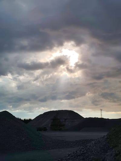 This is a medium-format, high-angle, vertical, color, exterior, daylight photograph of a gravel pit in Shalsky, Russia. The sky is filled with dark, heavy clouds that are partially broken in the center, allowing a bright shaft of sunlight to penetrate. In the foreground, on the left, is a large pile of dark gray gravel. To the right of that, a flat, gray gravel area extends back to a large, dark, conical pile of material, possibly coal or slag. In front of the large pile, two dark, heavy machinery vehicles are visible. Further to the right, another, smaller pile of dark material is visible, with a distant, lattice-work communication tower with red and white markings standing against the sky. The scene conveys a sense of an industrial or mining environment under a dramatic sky.