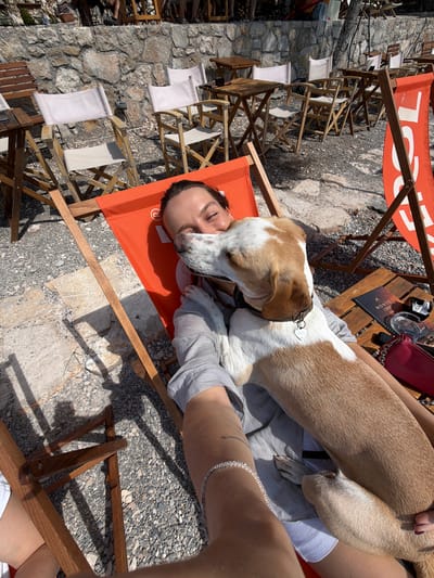 A woman is relaxing in an orange deck chair on a gravelly outdoor patio. She is lying on her back, with her head resting against the chair and her eyes closed. A brown and white dog is leaning over her, licking her face. The dog is wearing a black collar with a heart-shaped tag.
The patio appears to be part of a restaurant or bar, with several wooden tables and chairs visible in the background. The background also shows a stone wall and a large orange banner. The overall scene suggests a relaxed, outdoor setting, possibly a beachside establishment. The location is identified as Kuk, Montenegro.
