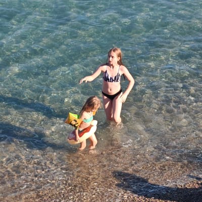 Two young girls are standing in shallow, clear ocean water. The girl in the foreground is smaller and wears a green bikini top with arm floaties that resemble donuts. She is holding a donut-shaped floatation device. The older girl behind her wears a black and white striped bikini. They are both looking towards the left side of the frame, with the older girl reaching out her left arm. The water is transparent, allowing the pebble-strewn seabed to be visible. Sunlight glints off the surface of the water, creating ripples and patterns. The scene appears to be a beach, likely during the daytime, given the bright lighting. The overall mood is one of playful enjoyment of the water.