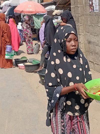 A woman wearing a black polka dot headscarf and colorful skirt holds a green bowl with red and green items. She is outdoors with other people and vendors' stands in the background. Some stands have umbrellas. The ground is light gray.