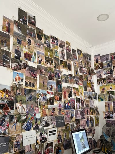 The image depicts an indoor room in Çiftlikköy, Turkey, with white walls almost entirely covered by a dense, overlapping collage of hundreds of printed photographs. These photographs, generally small to medium in size, primarily feature numerous individuals and groups, predominantly young adults, participating in various outdoor activities. A significant recurring theme is the presence of All-Terrain Vehicles (ATVs) or quad bikes, with subjects shown riding, posing alongside, or standing within natural, often hilly, landscapes. Many of the prints display curled edges and wear. Visible text on some photographs includes Arabic script and English phrases such as "WE HAVE CURRENCY EXCHANGE" and "One into Good". In the bottom right, a desk holds an open silver laptop displaying an image on its screen and a blue-bladed electric fan with a yellow base. A clear plastic water bottle with a blue cap is partially visible on the desk. The white ceiling features a single circular recessed light fixture. The walls exhibit plaster imperfections and discoloration, potentially from adhesive.
