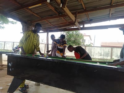 In Epe, Nigeria, a group of young men are playing pool outdoors under a corrugated metal roof supported by wooden beams. One man wears a yellow and green striped shirt, another a blue and yellow striped shirt, and another a black shirt with a red interior. The pool table is black with a green felt surface, holding multiple pool balls and cues. A low white wall runs in the background. A motorcycle with two riders is partially visible behind the wall.