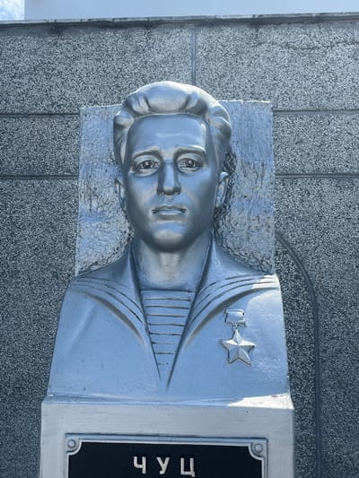 A silver bust of a young man in a sailor's uniform is mounted on a textured gray wall. The bust depicts the man from the chest up, with his hair styled neatly. He is wearing a collared shirt with horizontal stripes and a medal with a star attached to his chest. The bust is on a rectangular base, and below it, a black plaque with white Cyrillic lettering is partially visible, reading "ЧУЦ". The overall scene appears to be an outdoor monument. The location is the city of Takhtamukai, Russia.
