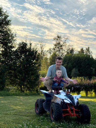 A man and a young boy are on a white and red quad bike in a grassy yard. The man is wearing a light green t-shirt and tan pants, and is crouching behind the boy. The boy is wearing a red and blue Spider-Man costume and is sitting on the quad bike. The quad bike has large, knobby tires and a blue seat. The background is filled with trees and a cloudy sky. The image is likely taken in a rural area, possibly in or near the city of Garevaya, Russia, given the type of vegetation and the overall atmosphere.