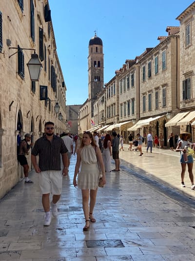 The image depicts a busy, sunlit street in Dubrovnik, Croatia, likely the Stradun, the main street of the Old Town. A young couple walks towards the viewer in the foreground. The man wears a black and white striped short-sleeved shirt and white shorts, with white sneakers and sunglasses. The woman wears a cream-colored sleeveless dress with ruffles and brown sandals. Behind them, a diverse crowd of tourists and locals stroll along the wide, paved street. The architecture is characteristic of Dubrovnik, with stone buildings lining both sides of the street, featuring arched windows, shuttered windows, and awnings above shop fronts. In the distance, a tall bell tower with a dark dome stands against a clear blue sky. The stone pavement is wet in places, suggesting recent rain or cleaning. The overall scene is one of vibrant daytime activity in a historic European city.