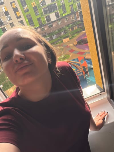 A young woman with light brown hair, wearing a dark red t-shirt, takes a selfie in front of a window. She is leaning against the windowsill with her right hand. Her nails are painted dark gray. She is wearing two rings on her right hand, one with a small clear stone. Through the window, a colorful courtyard with playground equipment and benches is visible. It is raining, with visible raindrops in the air and on the ground. In the background, a modern apartment building with green and cream colored facade and balconies is seen. The location is identified as Pervomaysky, Russia.