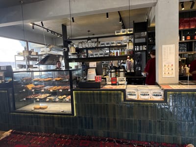 The image depicts the interior of a cafe, identified as "Submarine" and located in Krasnaya Polyana, Russia. The scene is captured from a frontal perspective, focusing on the serving counter and display area.
The serving counter features a dark green, vertically tiled backsplash illuminated by a strip of yellow LED lights along its top edge. On the counter, several white bags with "SUBMARINE" branding are displayed on a wooden shelf. To the left of these bags, a black electronic device, possibly a credit card reader, is visible. A portion of a cash register is also present. In front of the counter, a patterned red rug with a dark border lies on the floor.
The display case, situated to the left of the counter, showcases a variety of baked goods and desserts arranged on multiple shelves. These include cookies, pastries, and what appear to be chocolates or brownies. The display case is made of glass and has a black frame.
Behind the counter, a metal shelving unit is visible, holding various items, including bottles and packages. A person, partially obscured by the counter, is seen standing behind it, wearing a dark red shirt. Another individual, a woman, is seen in the background, also behind the counter, wearing a maroon garment and looking to the left. Further in the background, a kitchen exhaust hood is visible. The ceiling is made of concrete, with track lighting installed. A window with a view of an outdoor scene is present to the left. Several shelves are mounted on the wall behind the counter, displaying products and equipment.